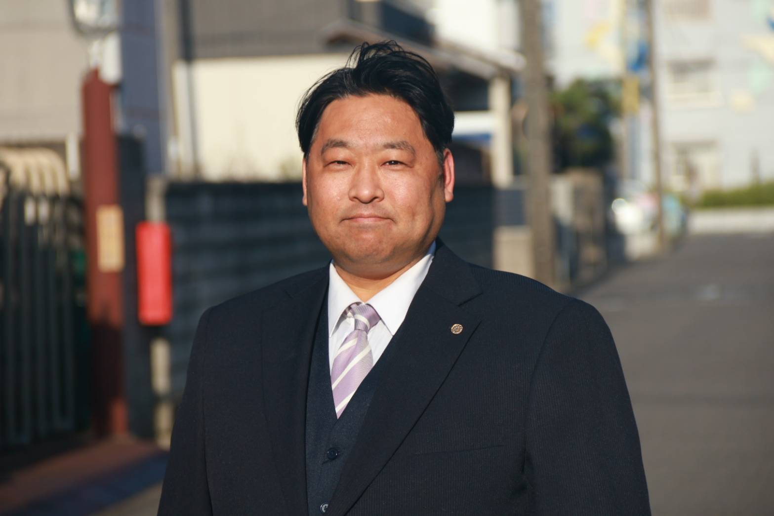 スーツ姿で正面を向いて立つ男性行政書士のプロフィール写真。背景には住宅街の道路と建物がやわらかくぼかされ、温かい日差しの中で穏やかな表情を浮かべている。